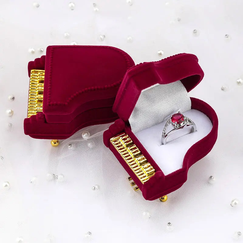 Burgundy velvet piano-shaped ring box with gold keys, open to show a ring inside