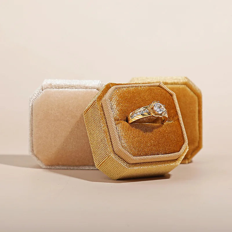 Tan octagonal velvet ring box open with a silver ring featuring a round stone
