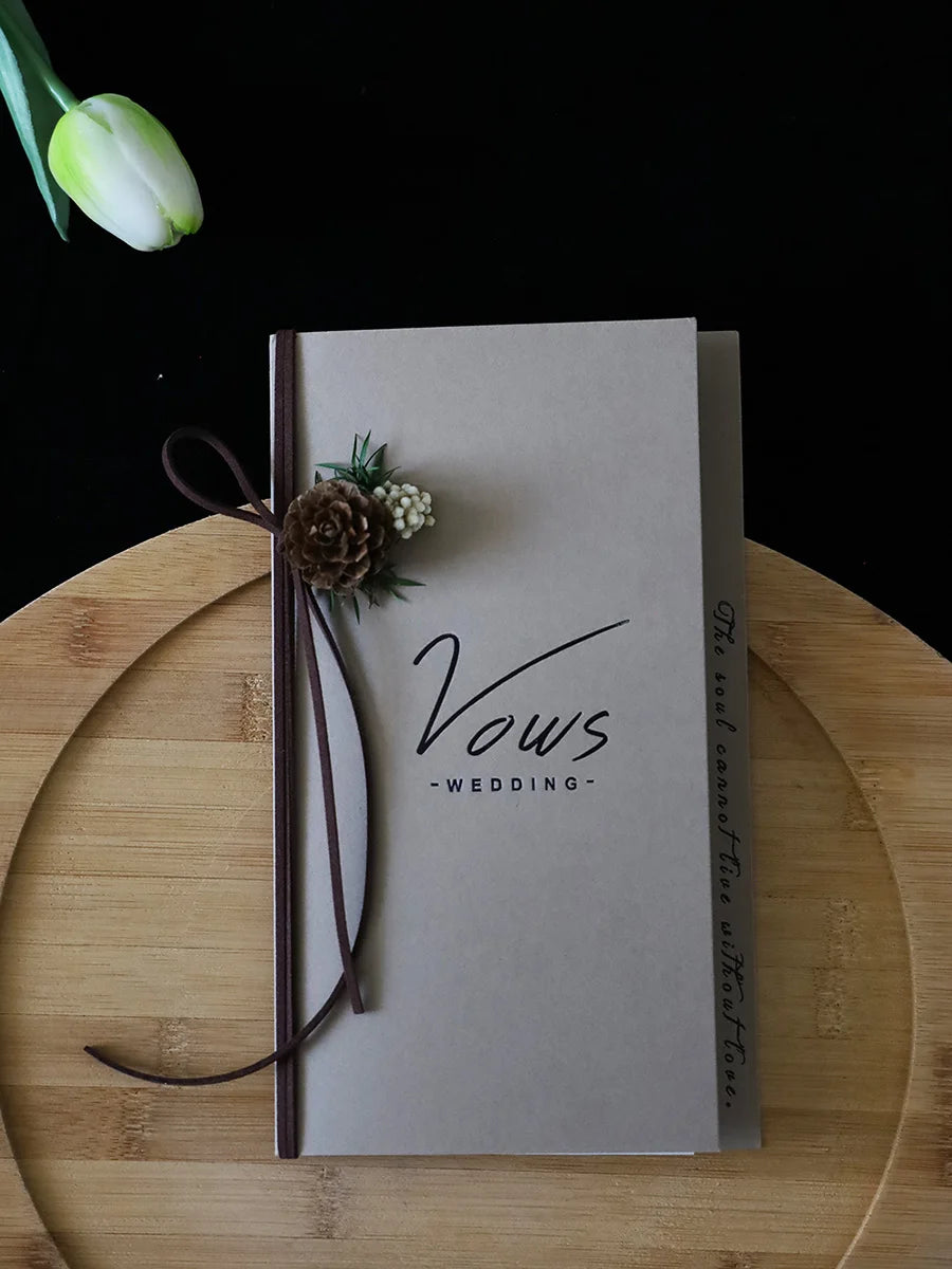 Ivory vows booklet with gold-foil “Vows Wedding,” dark ribbon wrap and dried flower cluster