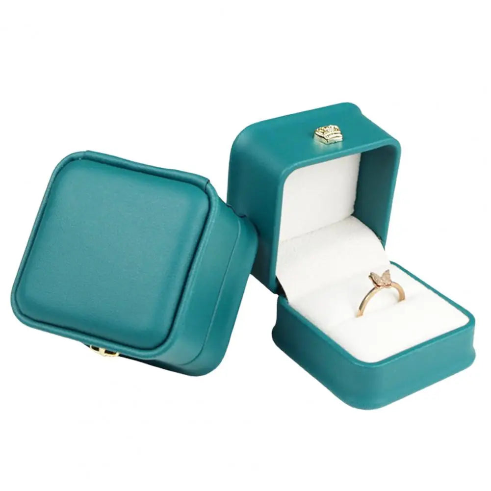 Teal green ring box with open lid, gold crown clasp, and ring on white cushion
