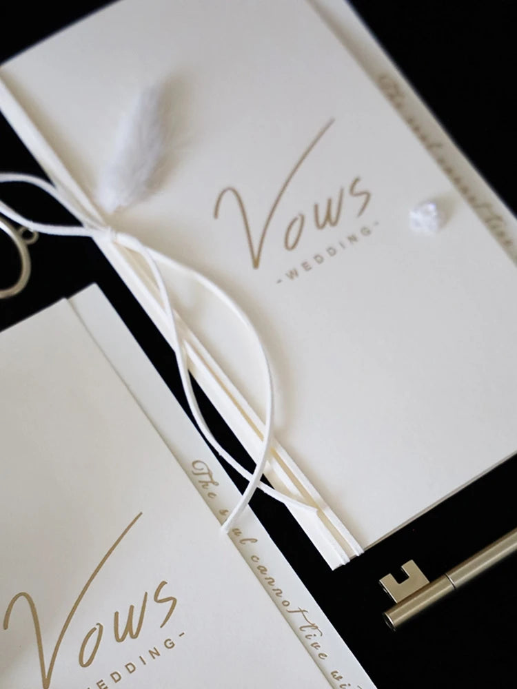 Close-up of ivory vow booklet cover with gold foil “Vows” and white ribbon tie