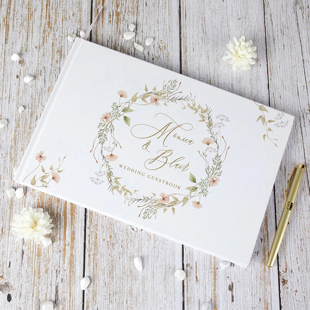 Wedding guest book with floral wreath cover and personalized names, tilted view, white with gold details