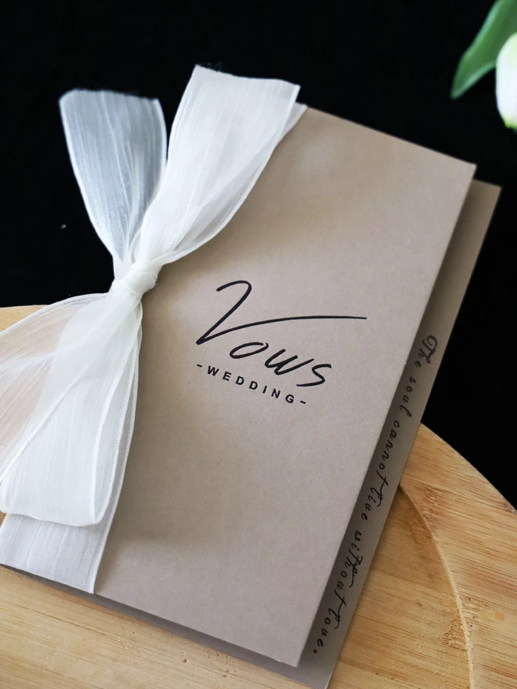 Ivory vows booklet with gold-foil “Vows Wedding” lettering, tied with white ribbon bow