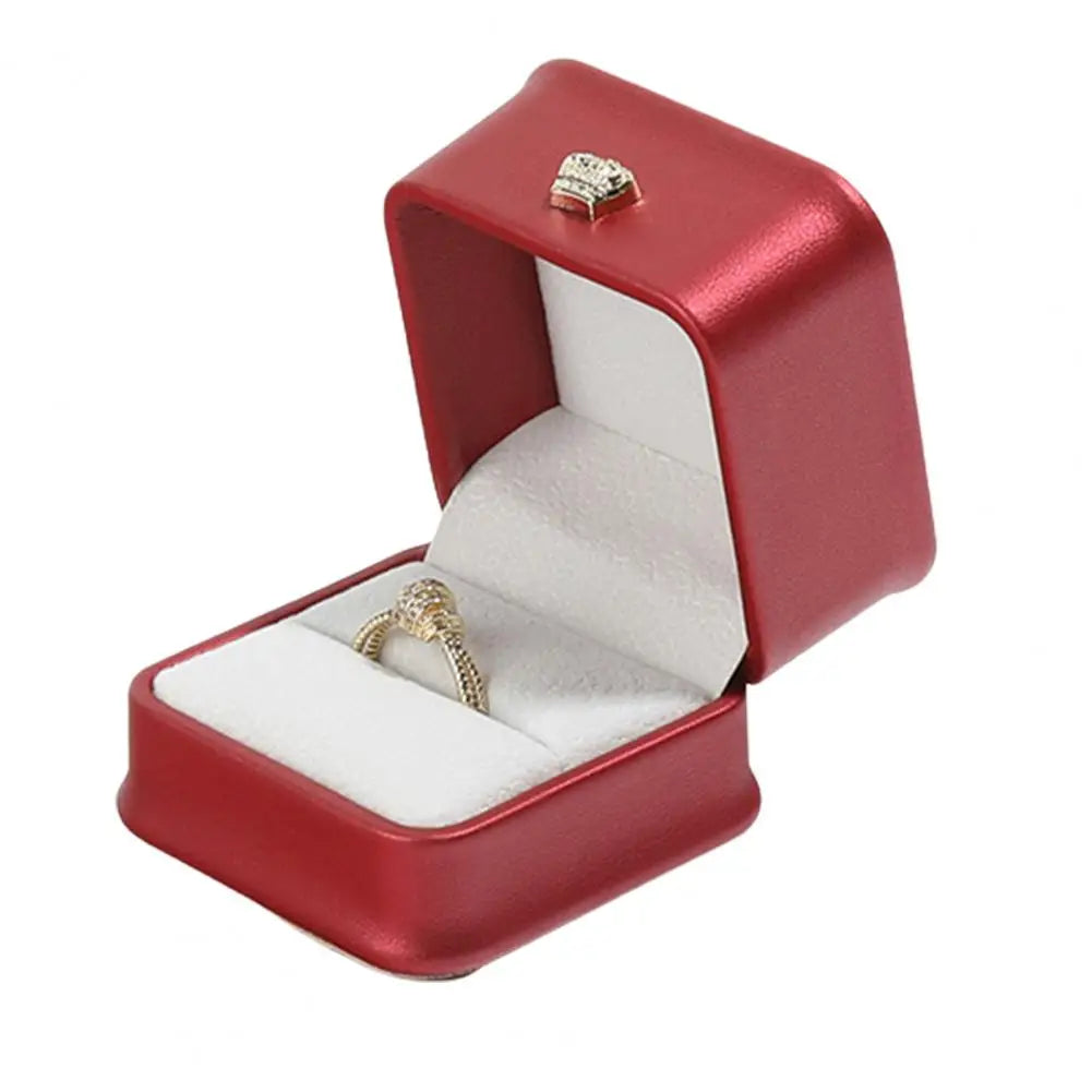 Red ring box with open lid, gold crown clasp, and ring on white cushion