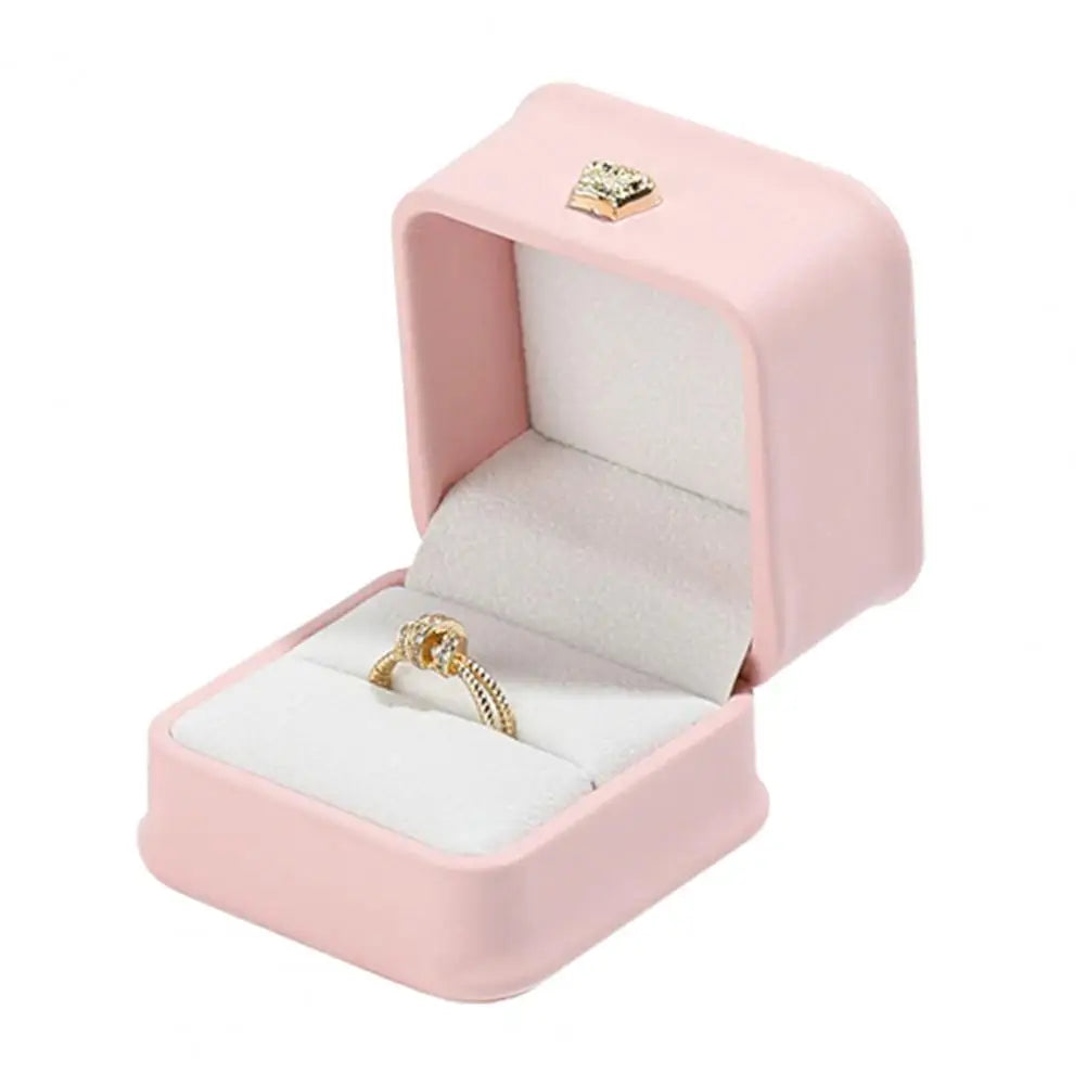 Pink ring box with crown clasp design, open with a gold ring on white cushion