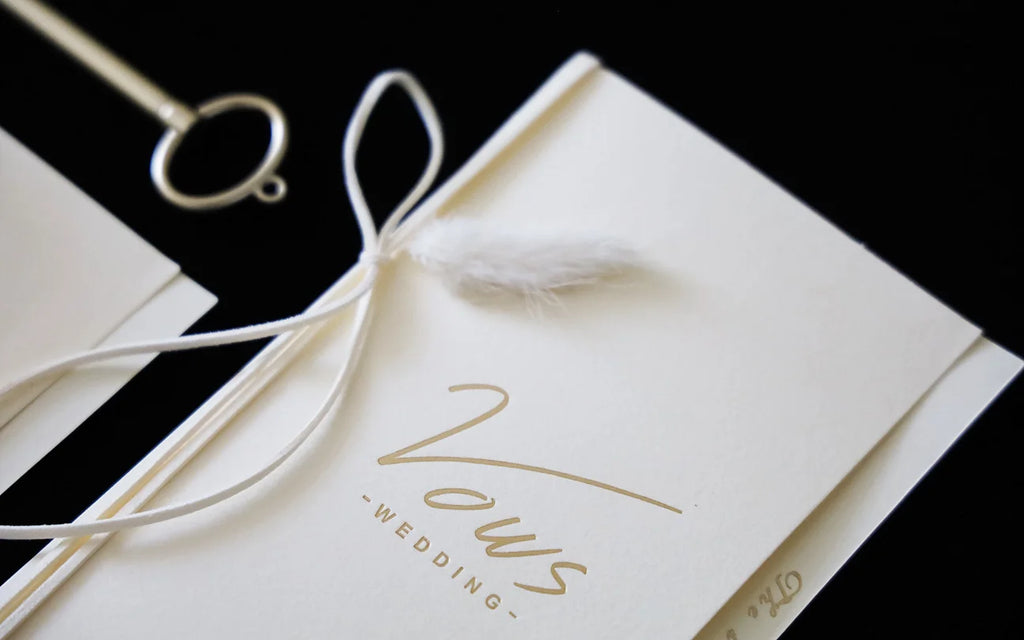 Close-up ivory wedding vow booklet with gold foil “Vows” and white ribbon bow