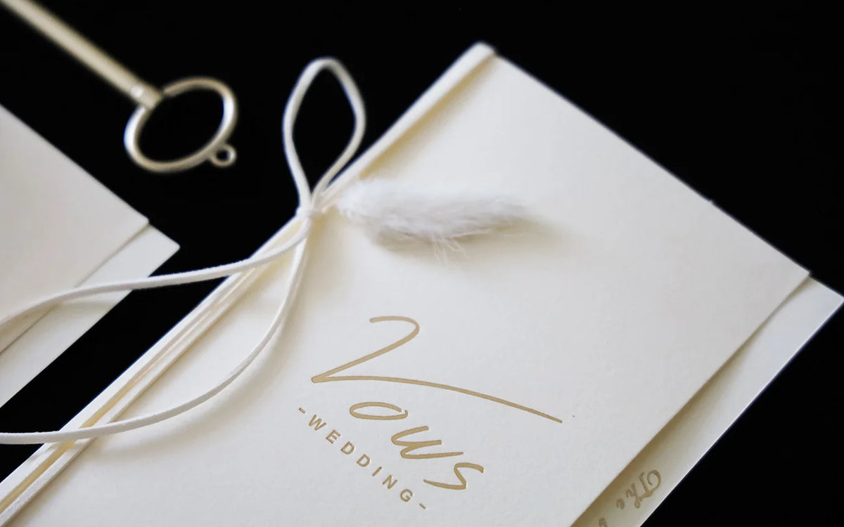 Close-up ivory wedding vow booklet with gold foil “Vows” and white ribbon bow
