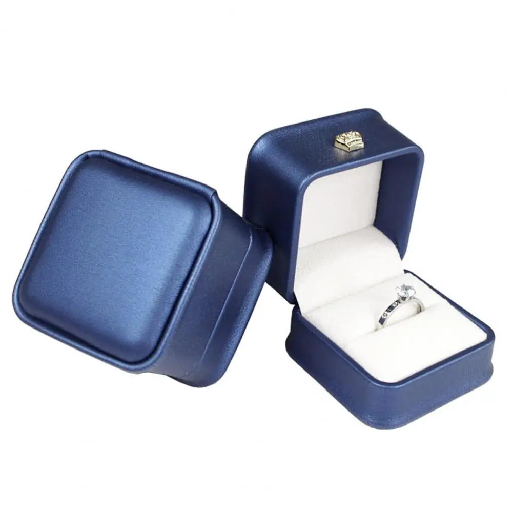 Navy blue ring box with open lid, gold crown clasp, and ring on white cushion