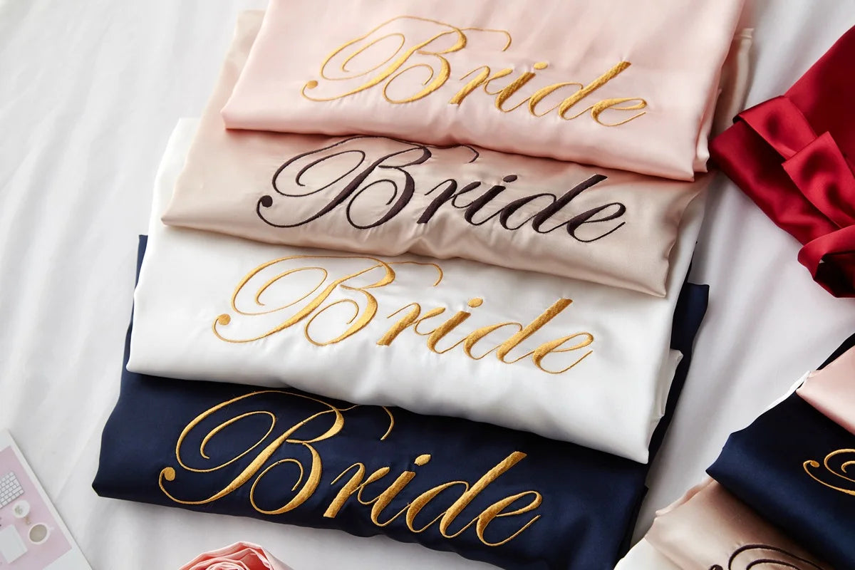 Satin bridal robes stacked, pale pink and navy with gold “Bride” script embroidery