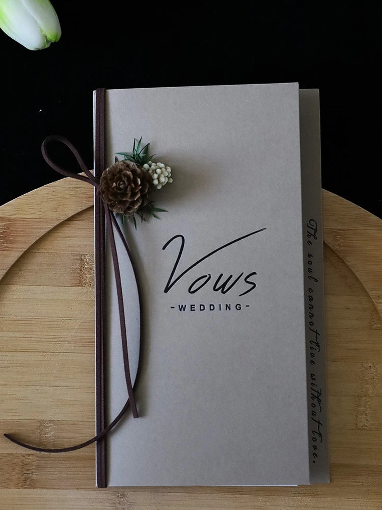 Ivory vows booklet with gold-foil “Vows Wedding” and dark ribbon with dried flower cluster