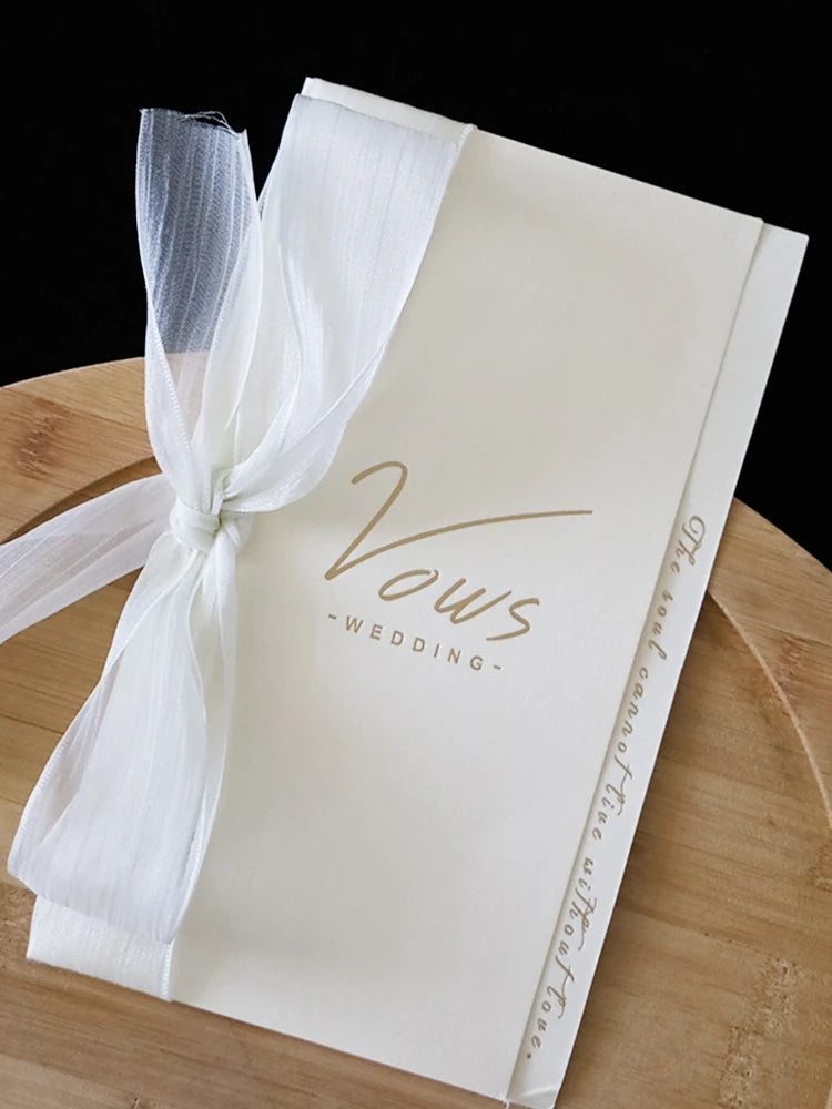 Ivory wedding vow booklet tied with long white ribbon, gold “Vows” lettering visible