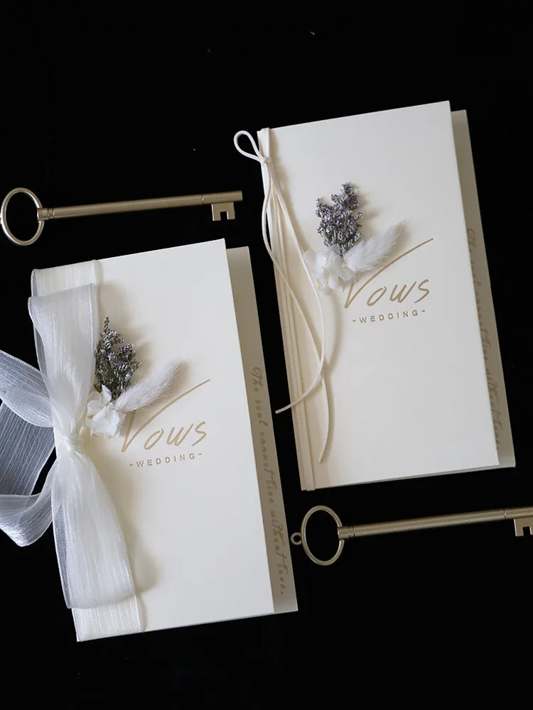 Ivory vows booklet set of two with gold foil lettering, floral detail, and tied white ribbon