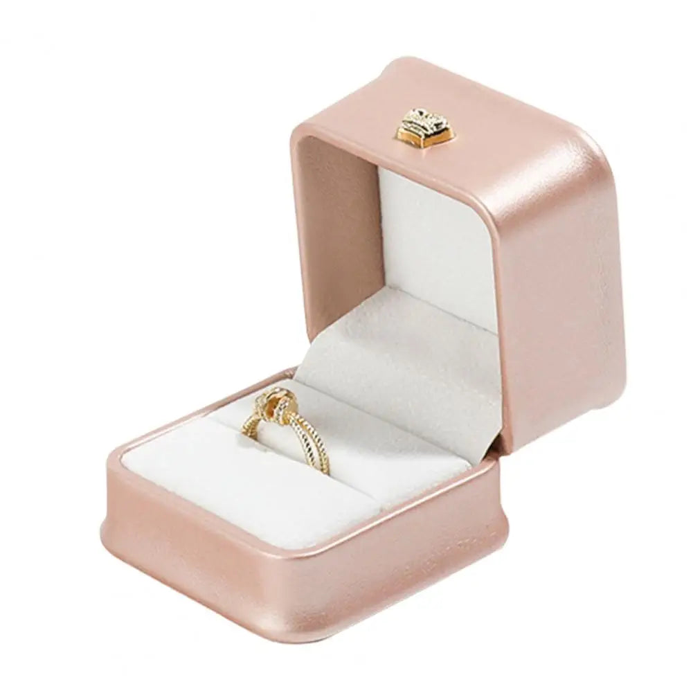 Blush pink ring box with open lid, gold crown clasp, and ring on white cushion