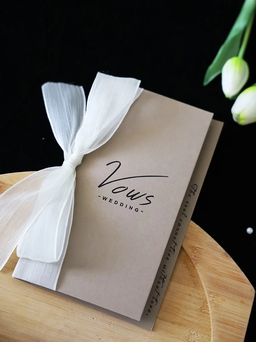 Ivory vows booklet with gold-foil “Vows Wedding” lettering, tied with flowing white ribbon