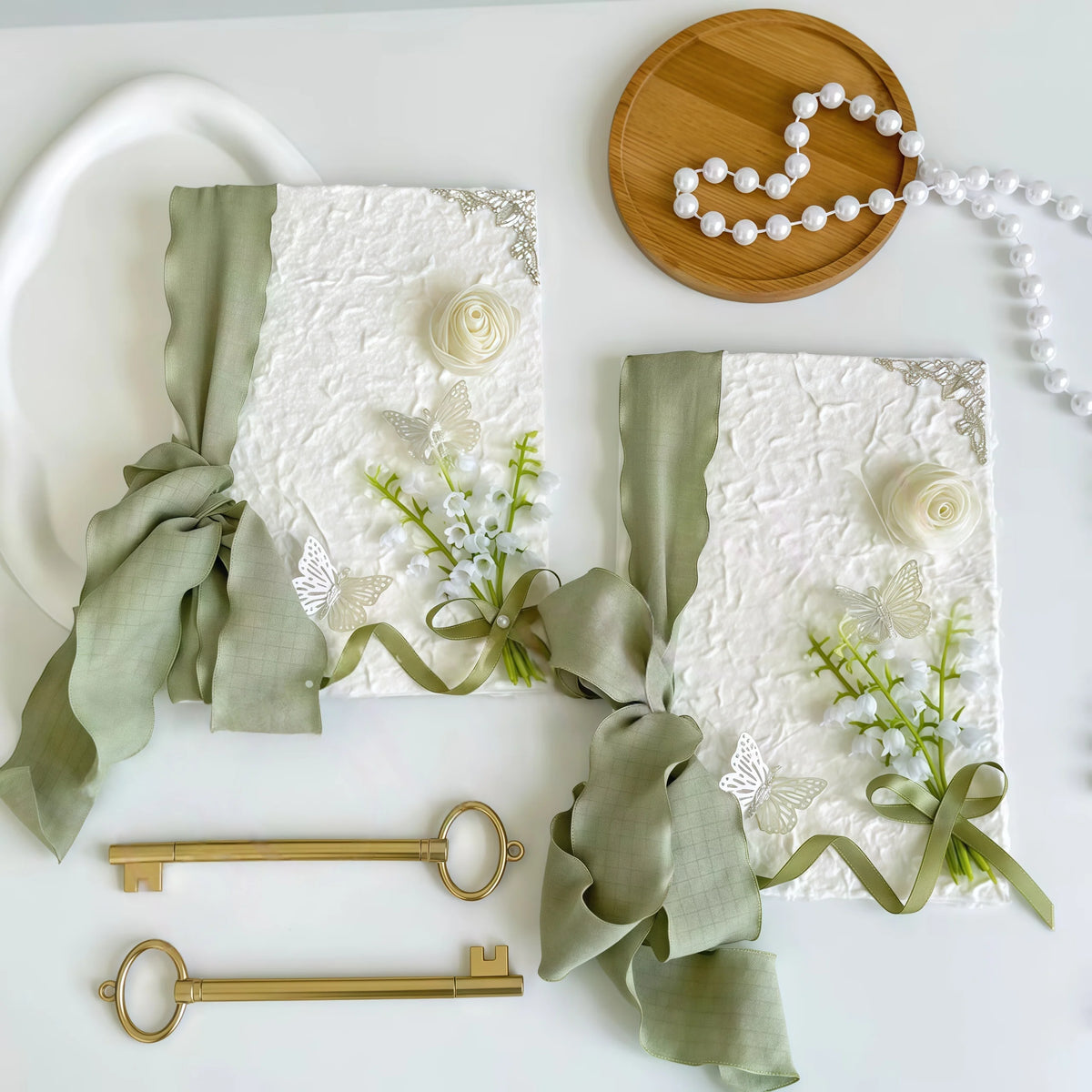 Two white wedding vow booklets with sage ribbon bows, rose detail, and small pearl clusters