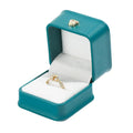 Teal ring box with open lid, gold crown clasp, and ring on white cushion