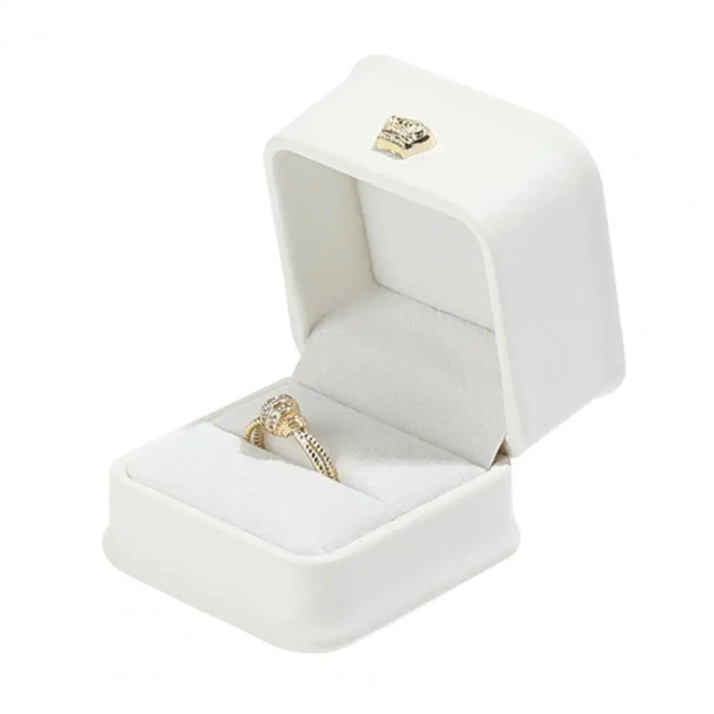 White ring box with open lid, gold crown clasp, and ring on white cushion